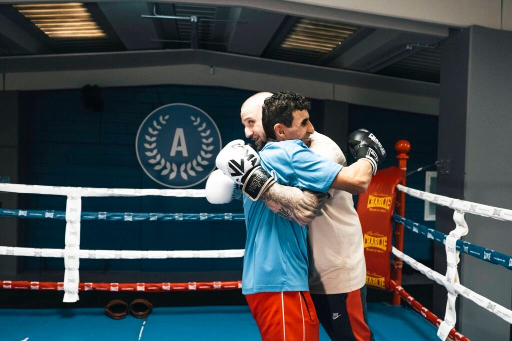 Dos hombres abrazándose dentro de un ring de boxeo durante un entrenamiento, uno de ellos con guantes y el otro con almohadillas de enfoque. Se aprecia el logo de Apolo Club Esportiu en la pared del fondo.