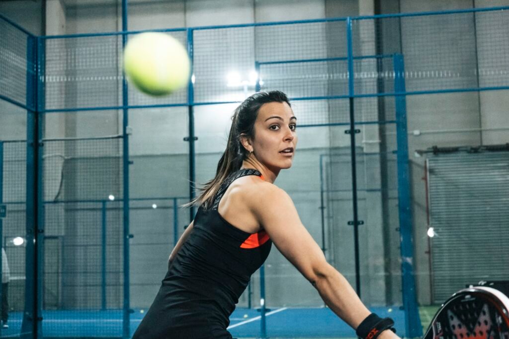 Mujer jugando pádel, con la pelota en pleno vuelo frente a su cara. Está concentrada en la pelota y se prepara para golpearla. Lleva ropa deportiva negra.