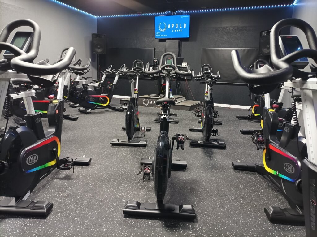 Sala de spinning con varias bicicletas estáticas dispuestas en filas, iluminada con luces LED y con una pantalla en la pared mostrando el logo de Apolo Gimnas.