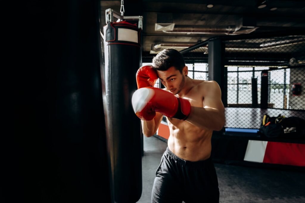 Un deportista sin camiseta, con guantes de boxeo rojos, se encuentra en posición de ataque mientras golpea un saco de boxeo en un gimnasio. La luz incide en su rostro y torso, destacando su concentración y fuerza.